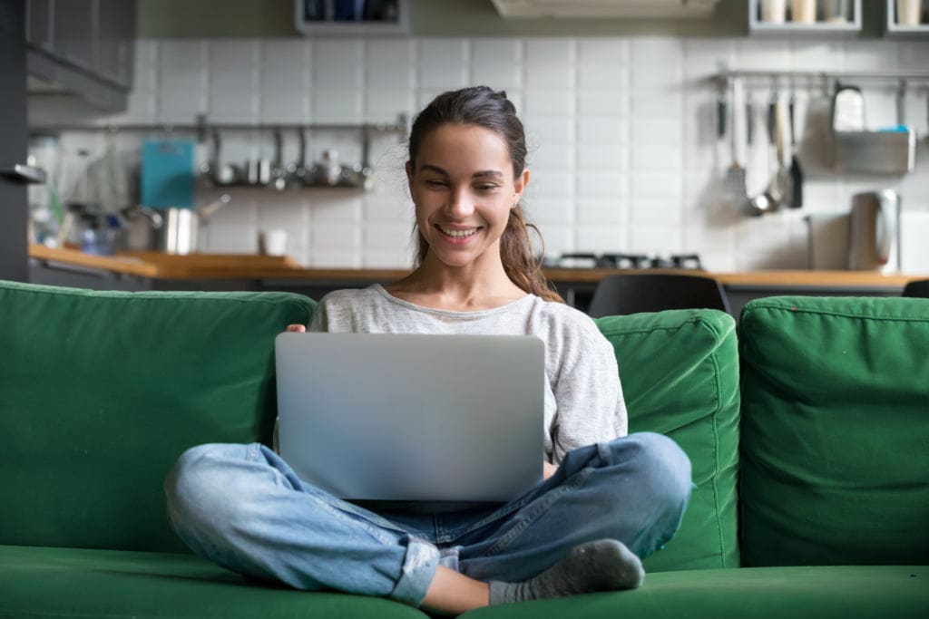 A smiling young woman is sitting on a couch with notebook computer on lap. A bright kitchen is in view behind the woman and couch.